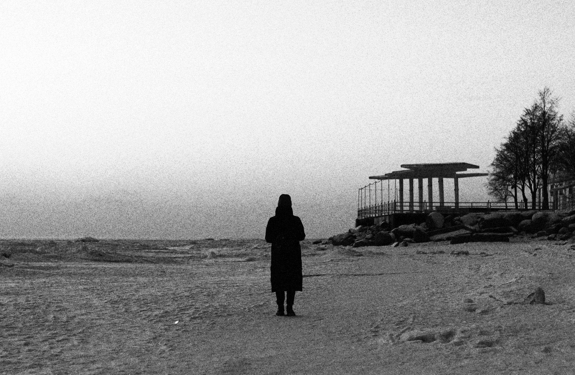 Mysterious silhouette of a woman in winter coat on a deserted beach with a distant structure.