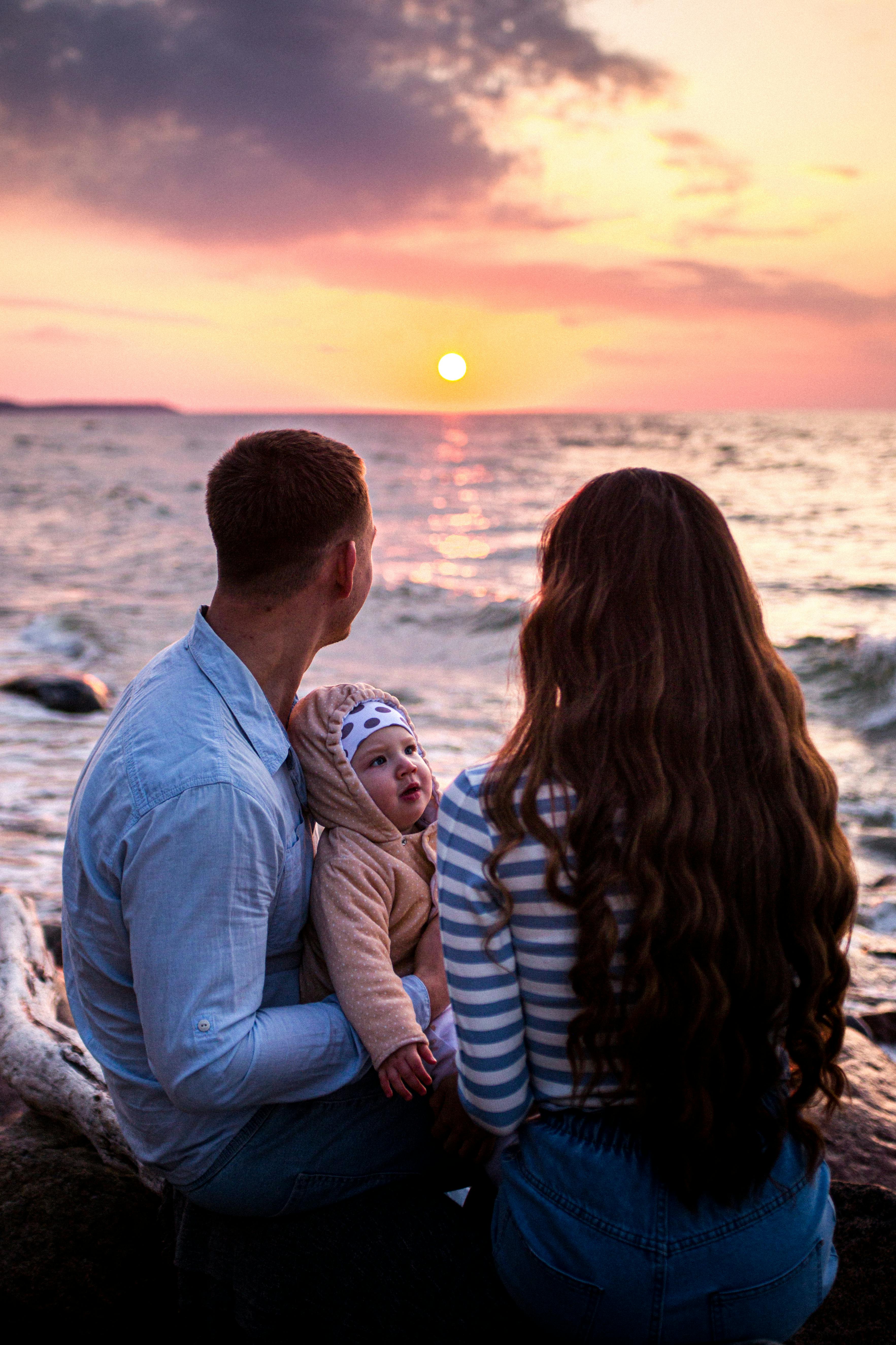 couple with baby sitting rock near body of water during sunset