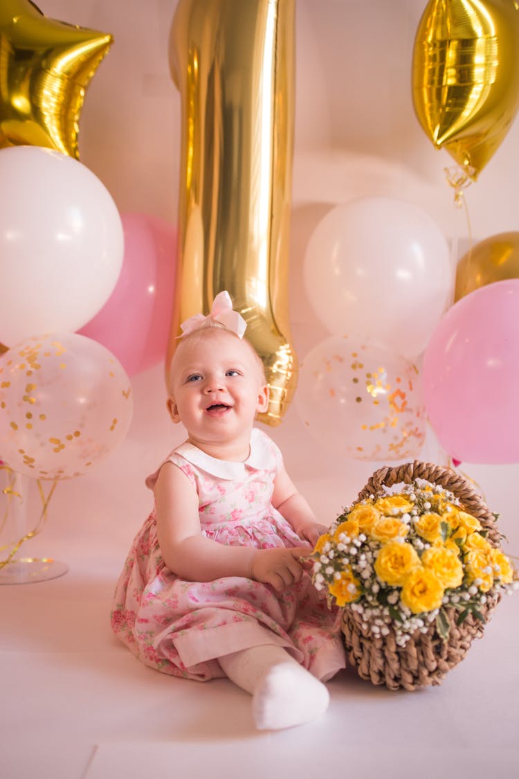 Baby Girl In Pink Dress Sitting On Floor With Basket Of Flowers
