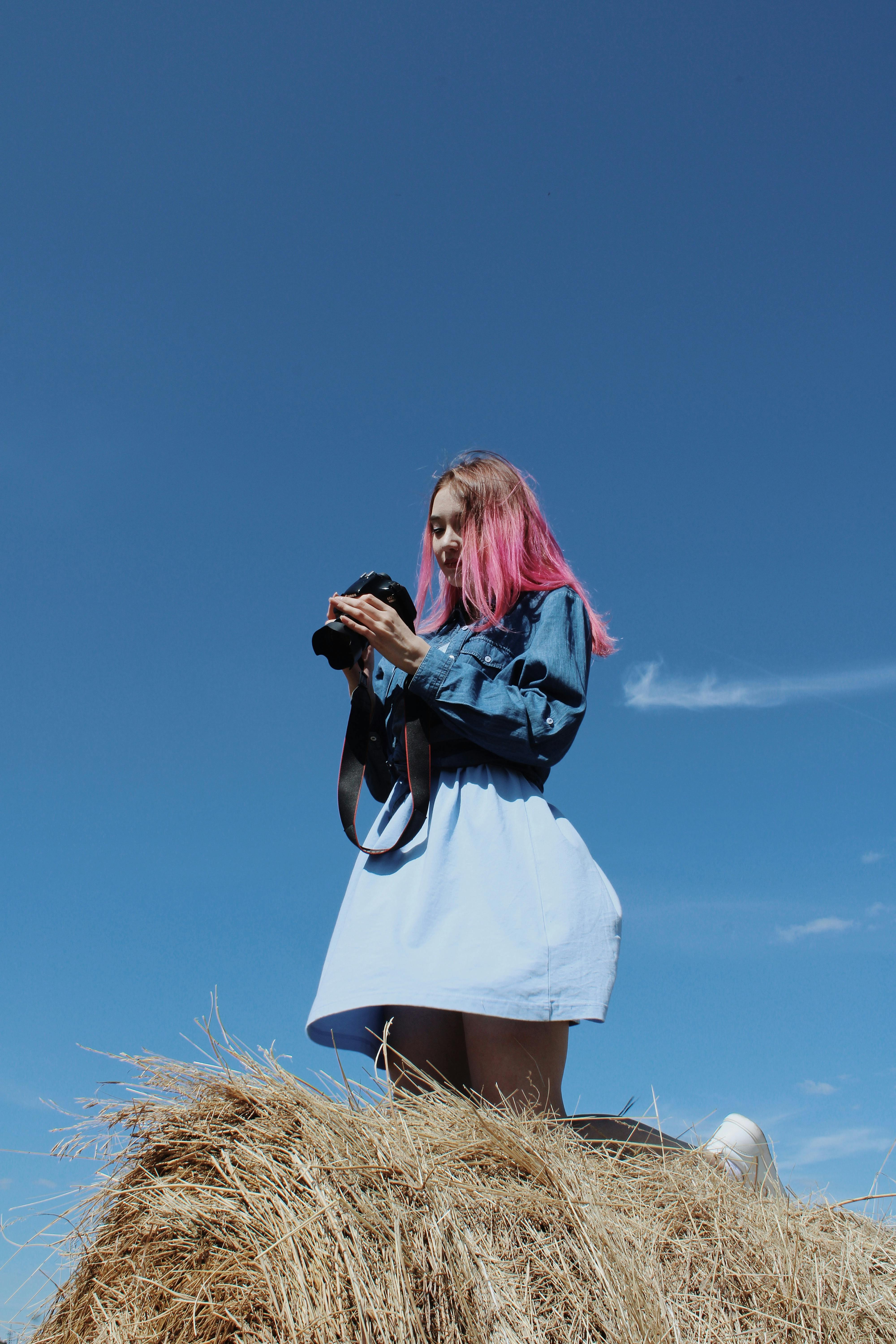 A woman with pink hair holding a camera stands on a haystack against a clear blue sky.