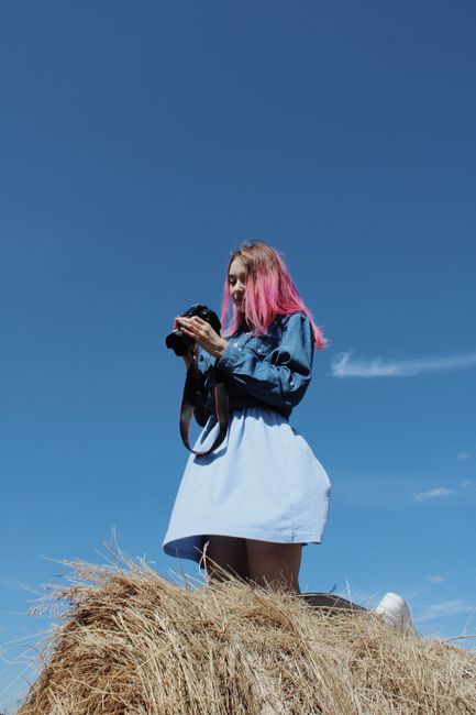 A woman with pink hair holding a camera stands on a haystack against a clear blue sky.