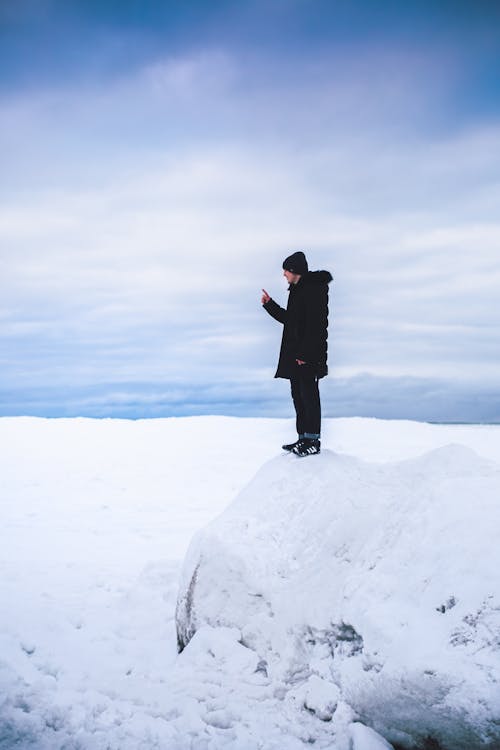 Man in Black Puffer Jacket Standing on Snow-Covered Rock under the Cloudy Sky