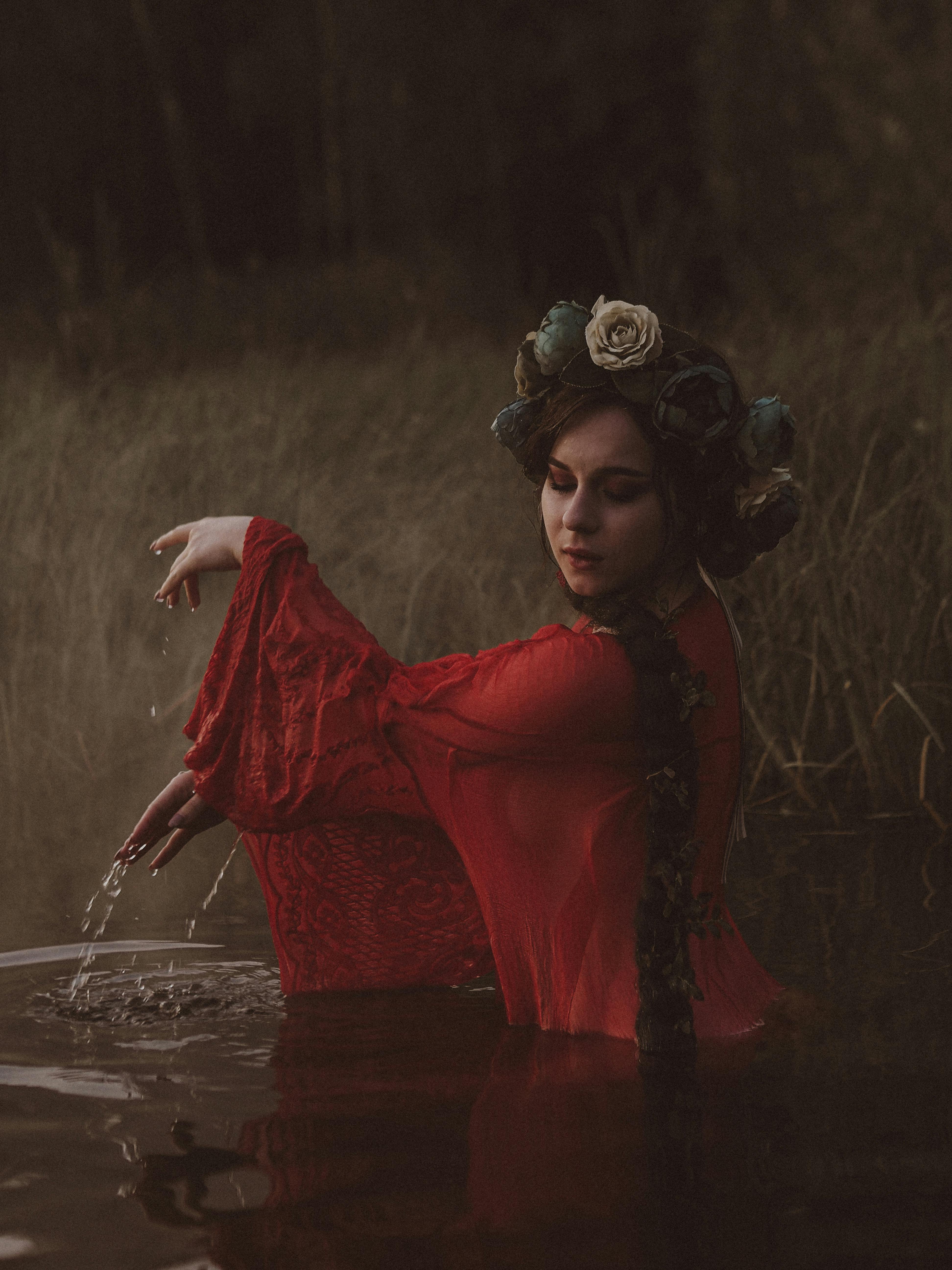 A woman in a red dress with floral headwear poses by a serene water landscape.