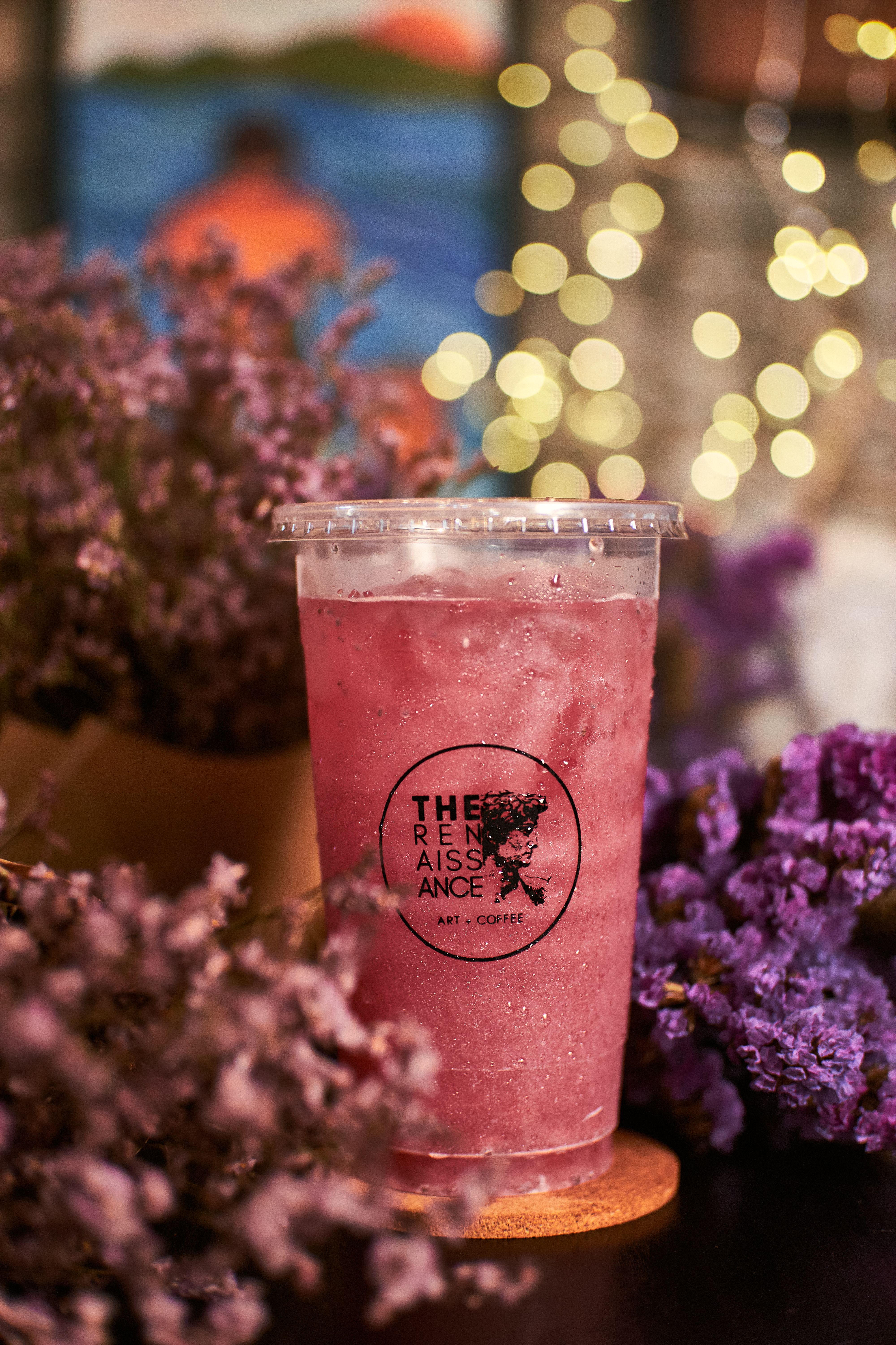 A refreshing iced pink drink in a plastic cup surrounded by vibrant purple flowers and bokeh lights.