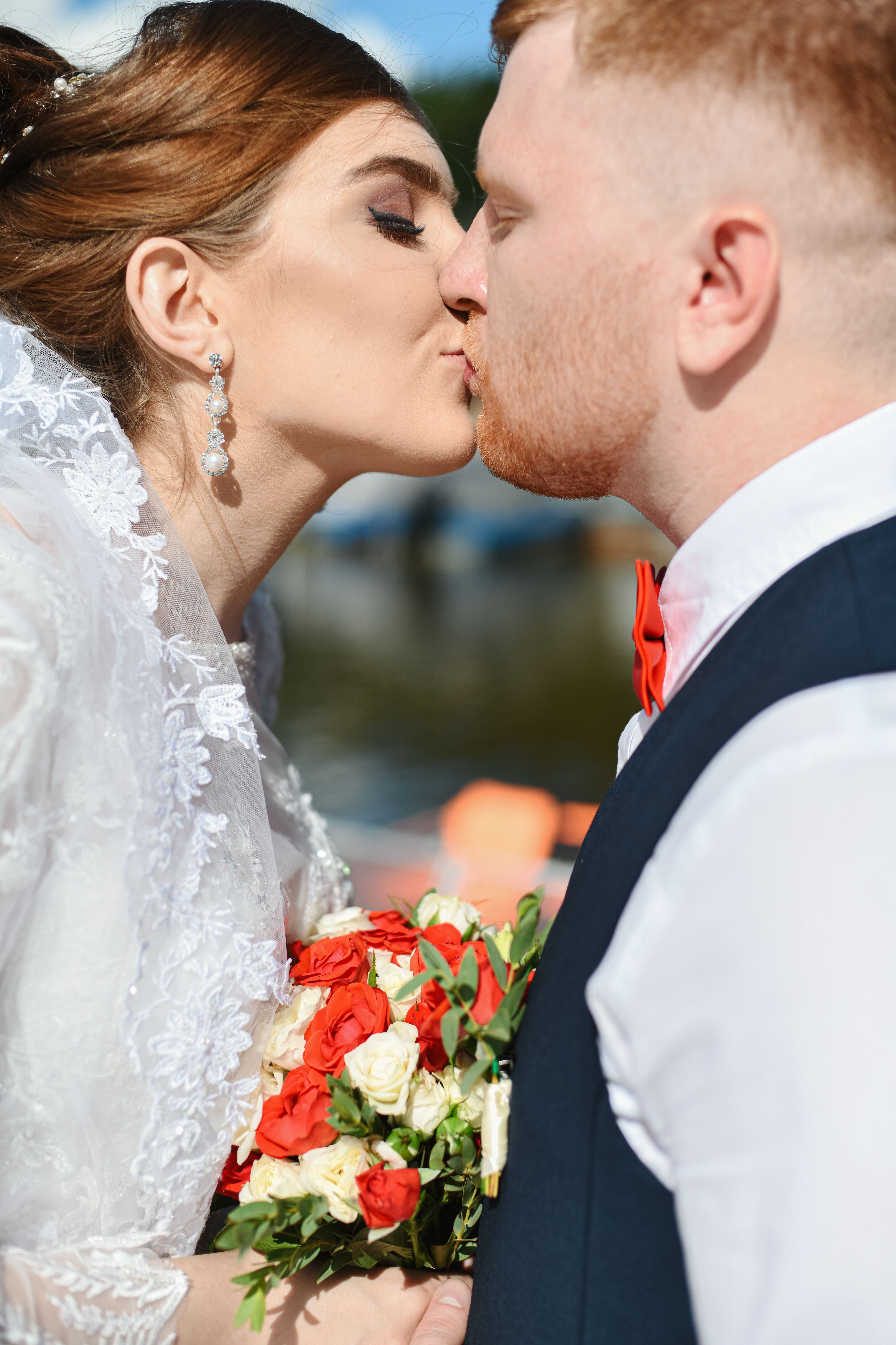 Bride and groom share a loving kiss outdoors, holding a red and white floral bouquet.