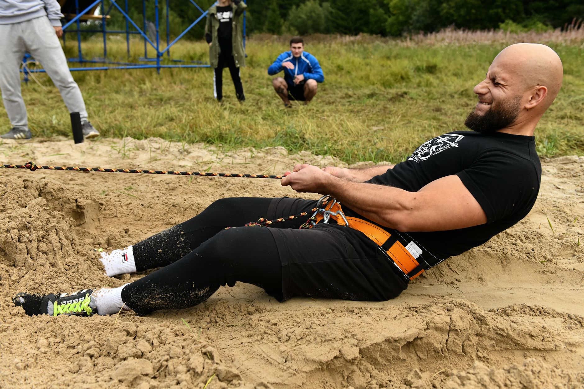A bald, bearded man using harness and rope for strength training in sand outdoors.