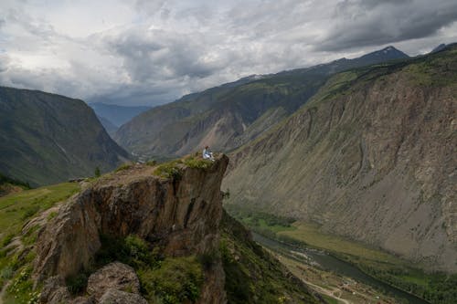 Person Sitting on Cliff Edge Under Gray Sky