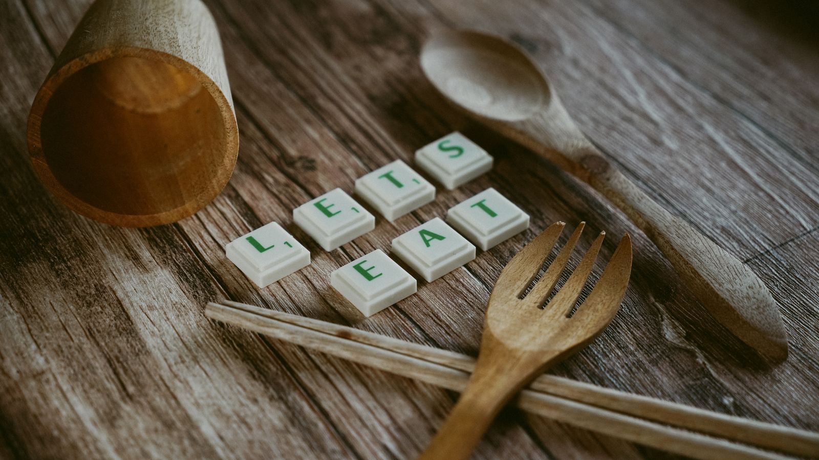 A rustic table setting featuring wooden utensils and tiles spelling 'LET'S EAT' on a wooden surface.