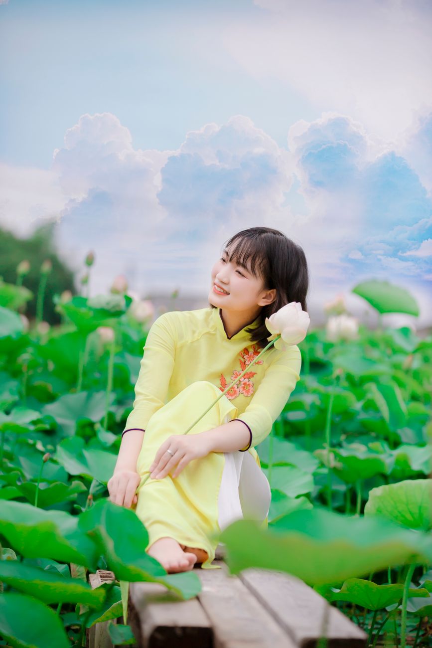 Young woman in traditional yellow ao dai sitting among lotus flowers under a bright sky.