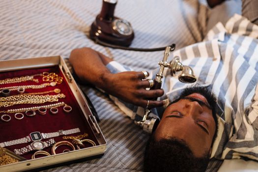 Bearded man lying on bed using antique phone with jewelry set nearby.