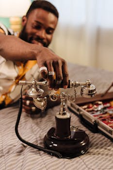 A joyful man adjusts a vintage rotary phone, surrounded by luxury jewelry on a bed.