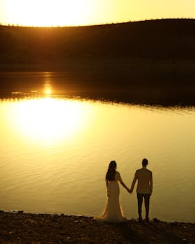 A couple stands holding hands at sunset by a serene lake, capturing a romantic wedding moment.
