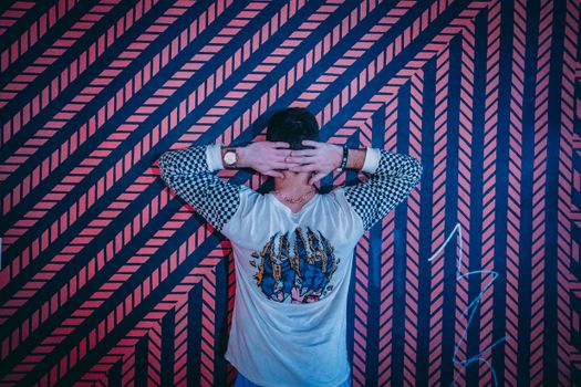 Man stands with hands on head in front of a vibrant geometric patterned wall, showcasing urban style.