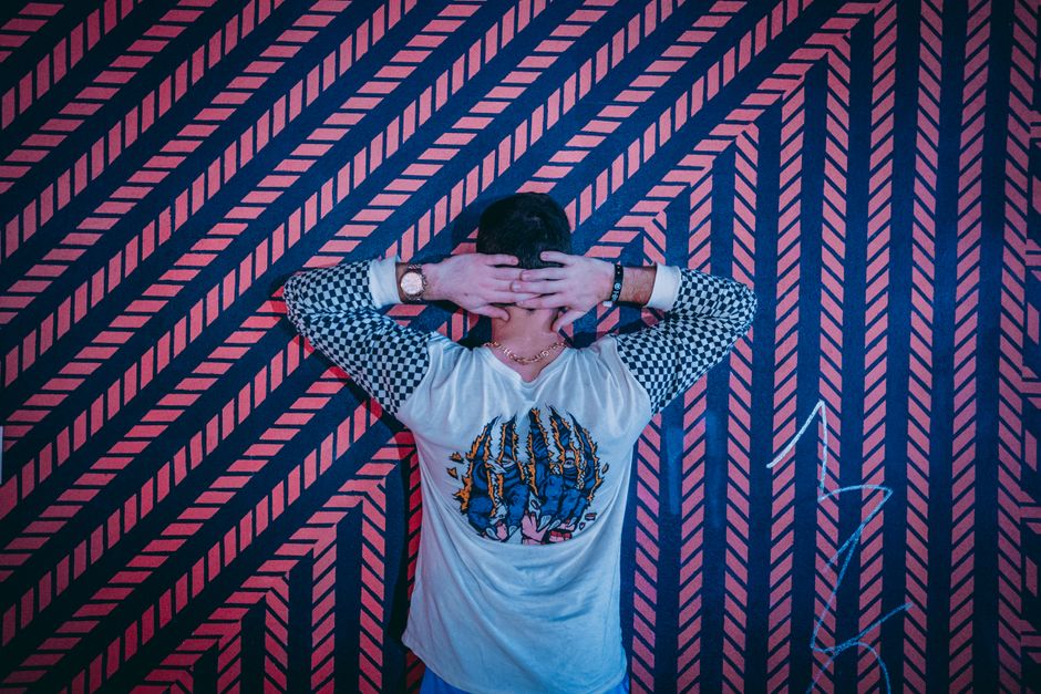 Man stands with hands on head in front of a vibrant geometric patterned wall, showcasing urban style.