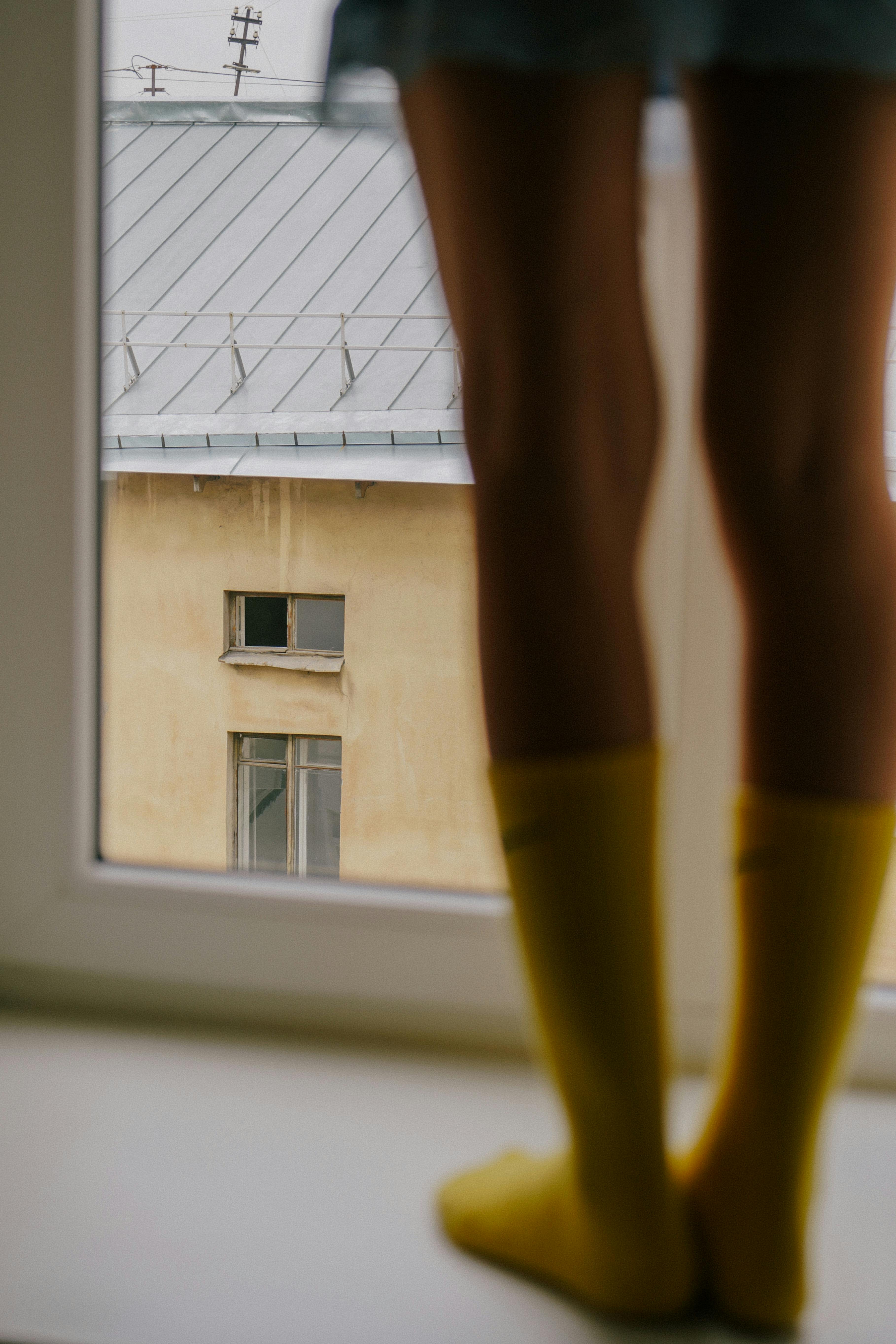 Close-up view of a person standing by a window wearing yellow socks, looking out at a building.