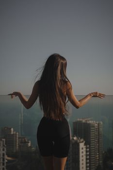 A woman with long hair stands on a balcony, overlooking a cityscape through a glass railing.