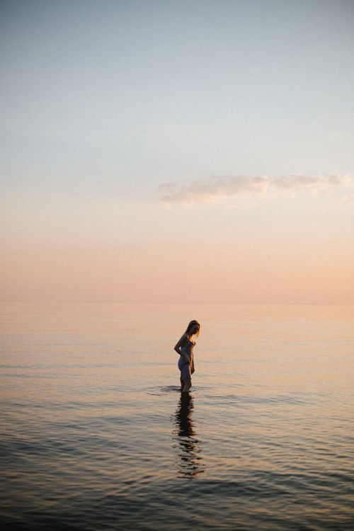 Free A woman in a dress stands peacefully in the ocean at twilight, enjoying the serene, pastel horizon. Stock Photo