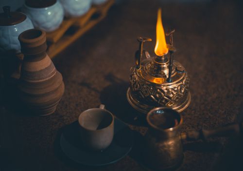Free A cozy and nostalgic coffee setup featuring an oil lamp, vintage pots, and cups on a warm-toned surface. Stock Photo