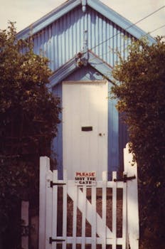A quaint vintage house entrance with a white gate and door, surrounded by greenery.