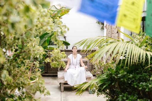A woman practices meditation on a wooden bench surrounded by vibrant garden plants.