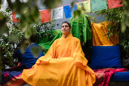 A peaceful woman meditates outdoors wearing vibrant Buddhist robes, surrounded by prayer flags and greenery.