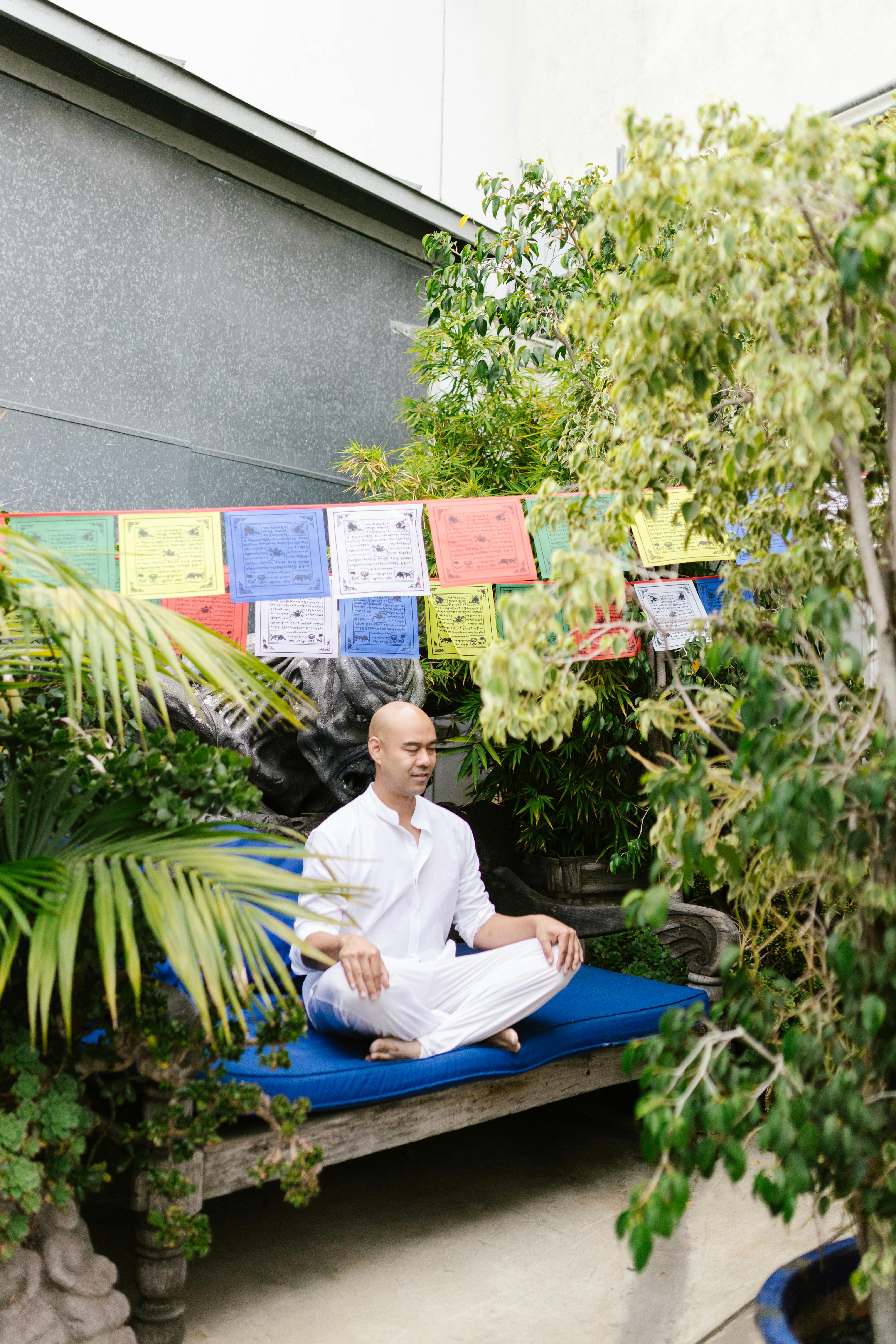 A man in white clothes meditates peacefully outdoors among lush greenery and colorful prayer flags.