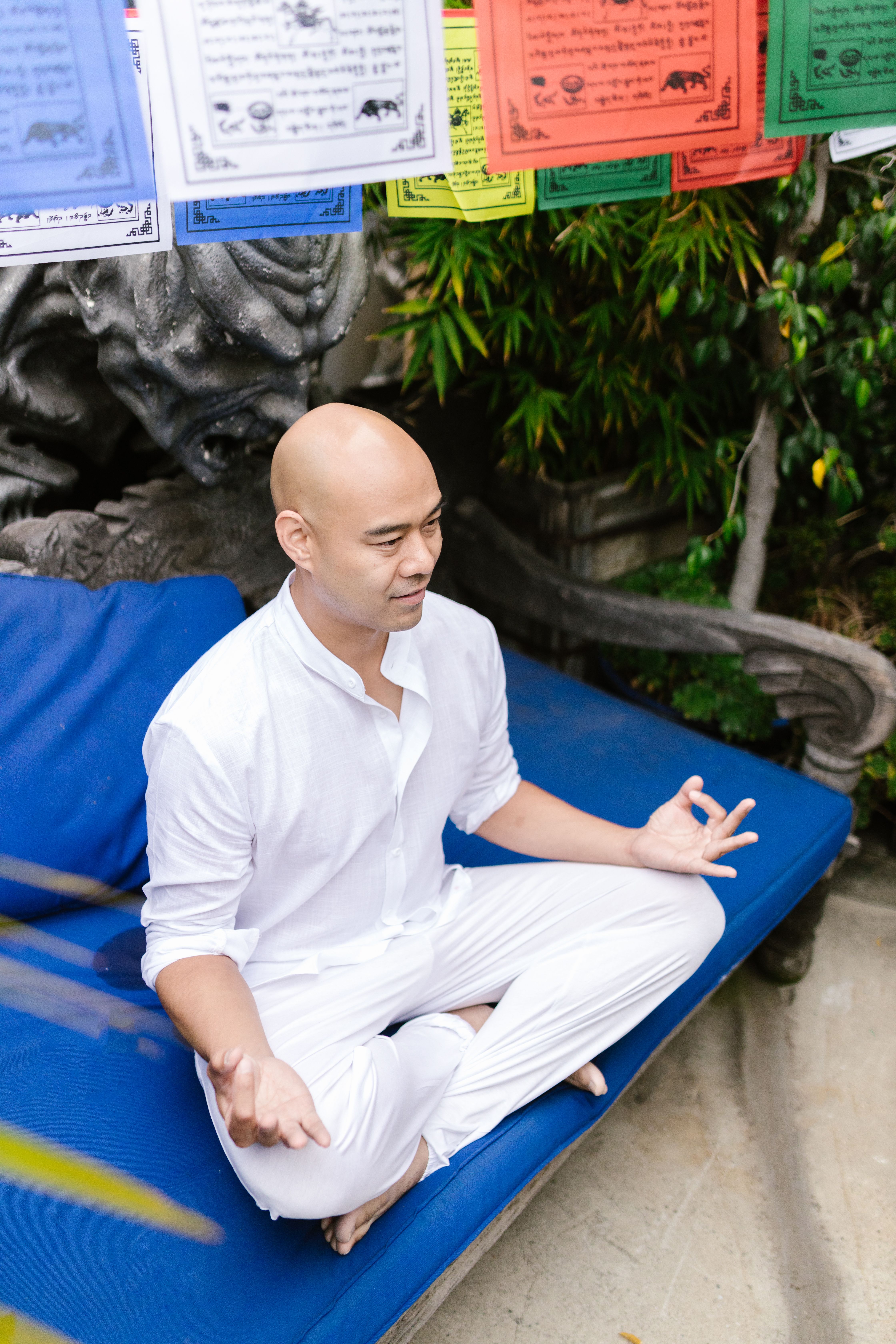 A man in white meditates peacefully outdoors surrounded by nature and vibrant flags.