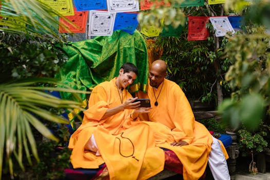 Two monks in bright robes share a moment with smartphone, surrounded by lush greenery and colorful prayer flags.