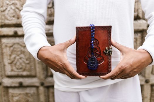 Hands holding a decorative book with a Buddha design and blue beaded necklace.
