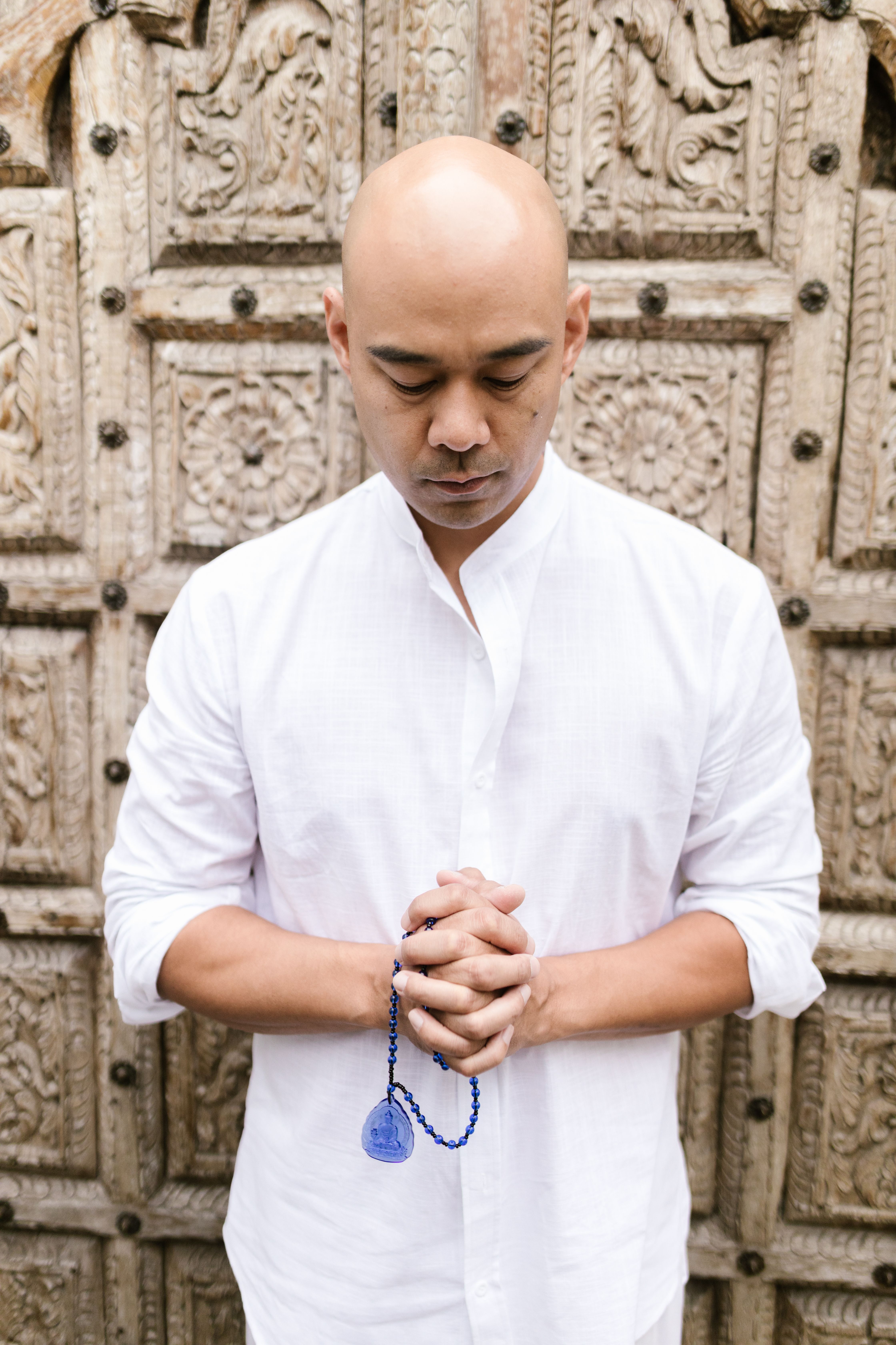 Bald man in white shirt meditates holding blue prayer beads before rustic wooden door.
