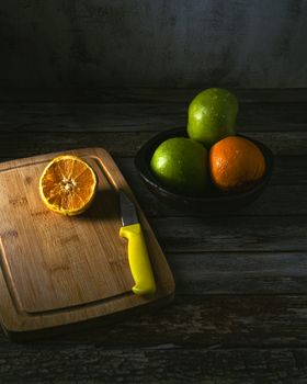 A rustic scene with a half orange, green apples, and a knife on a wooden board.