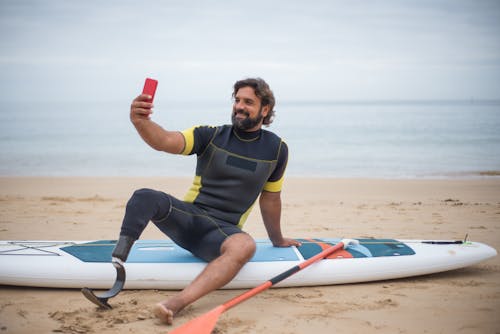 Man in Wetsuit Sitting on Surfboard while Taking Picture of Himself using His Cellphone
