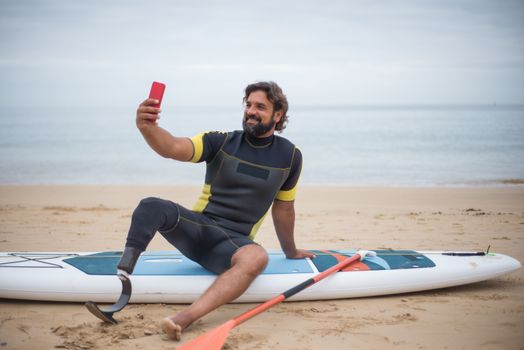 A man in a wetsuit with a prosthetic leg takes a selfie while sitting on a surfboard at the beach.