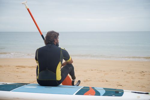 Free A man with a prosthetic leg sits on a paddleboard at the beach, enjoying a calm ocean view. Stock Photo