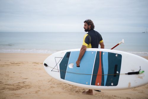 Free Bearded man holding a paddleboard on a sandy beach, ready for water sports in Portugal. Stock Photo