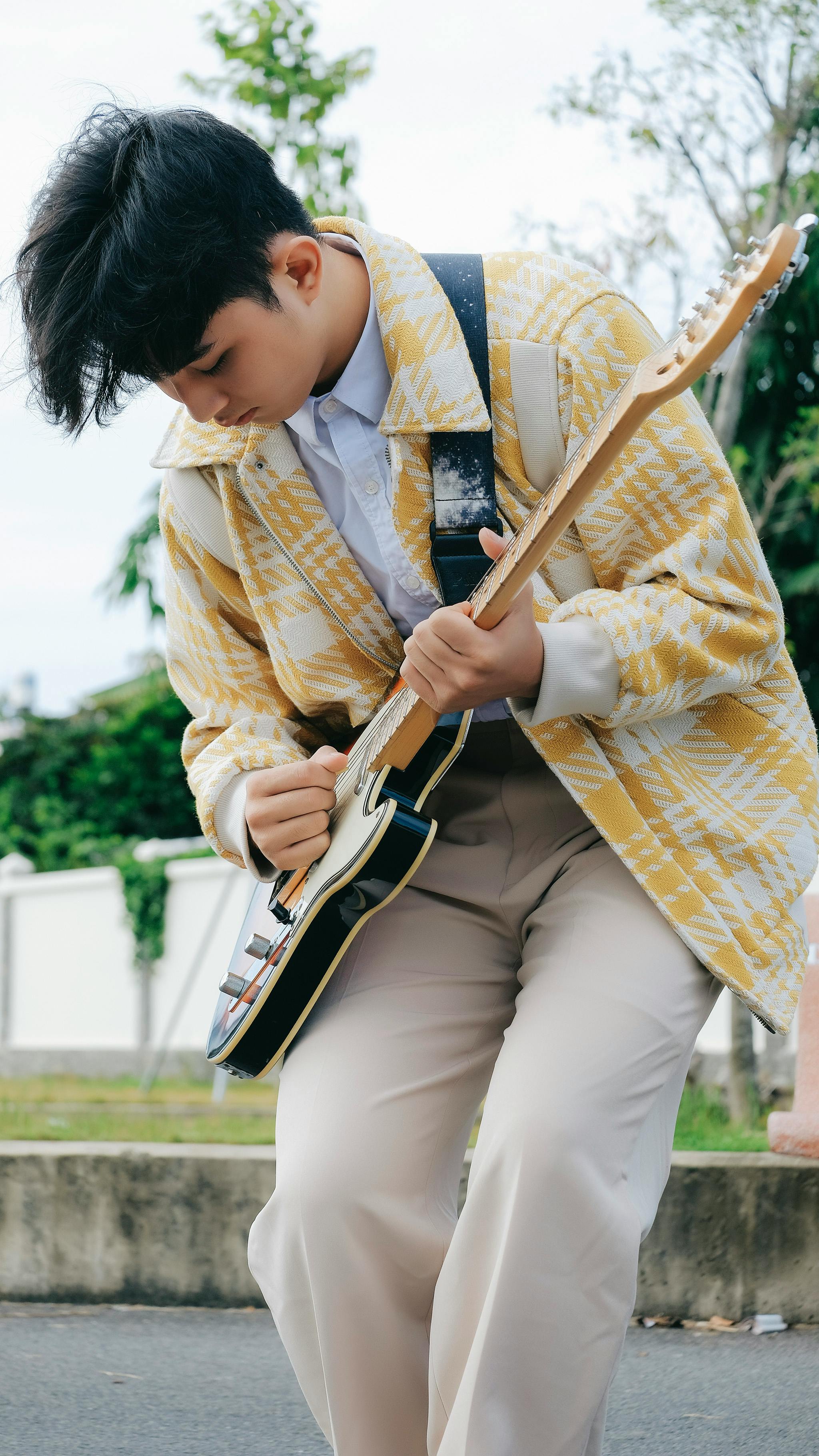 A young musician playing an electric guitar in a stylish yellow jacket outdoors.
