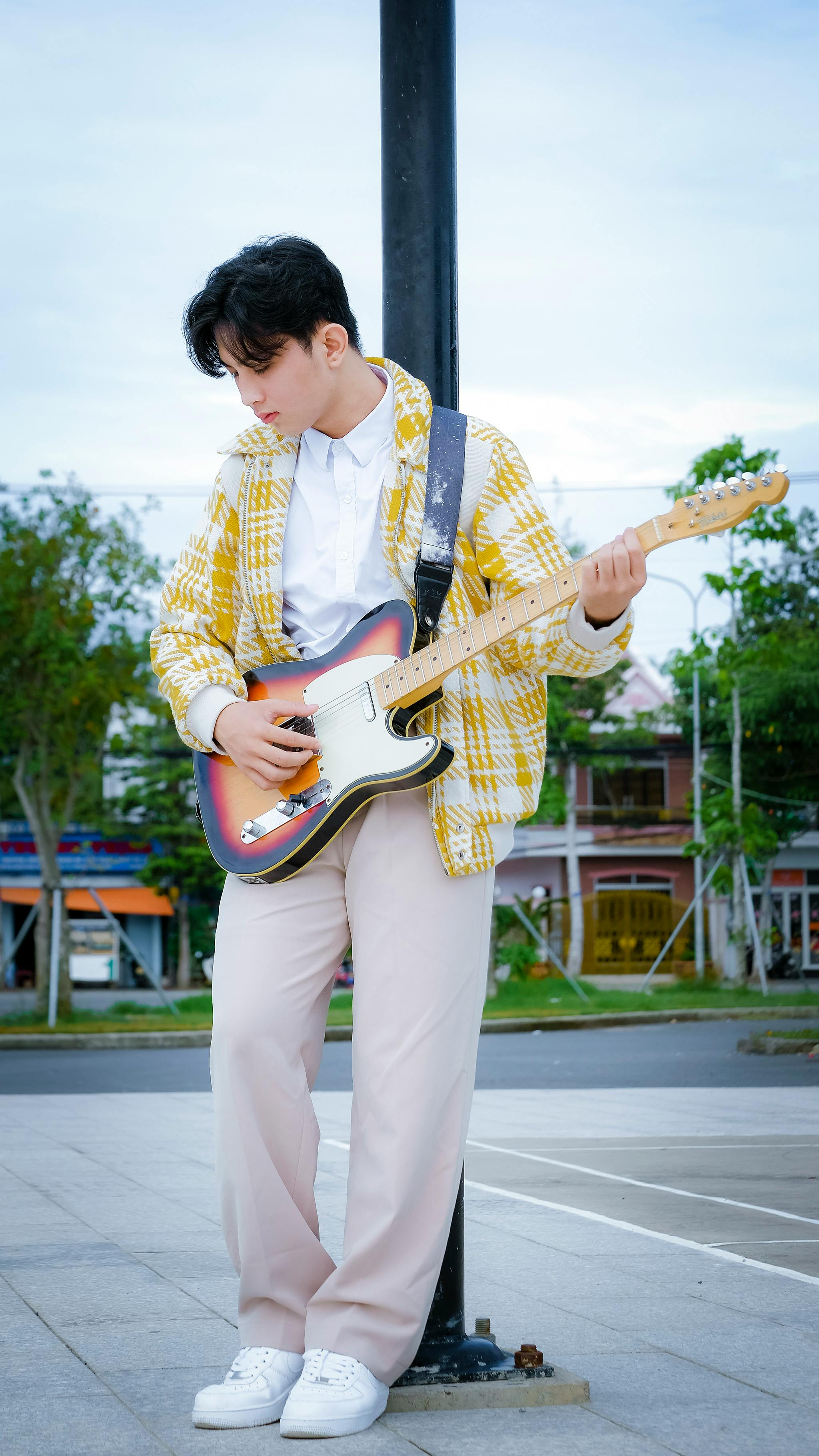 A young man in a plaid jacket playing guitar outdoors in a Vietnamese city.