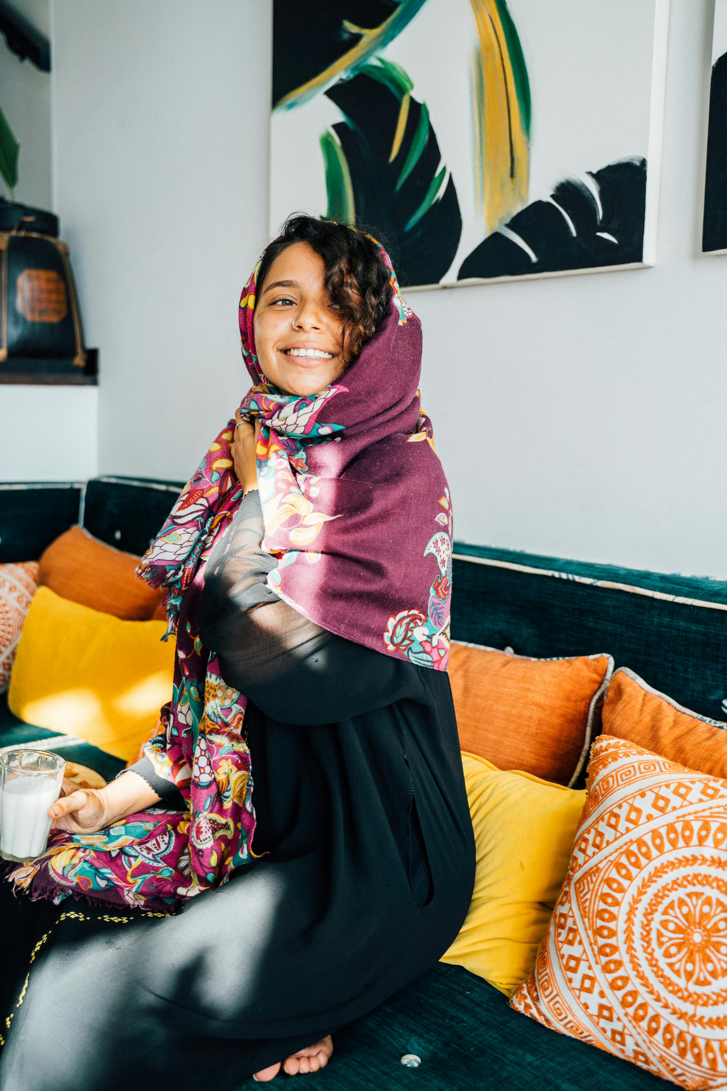 A cheerful woman in a colorful hijab sits on a vibrant couch, enjoying a relaxing day indoors.
