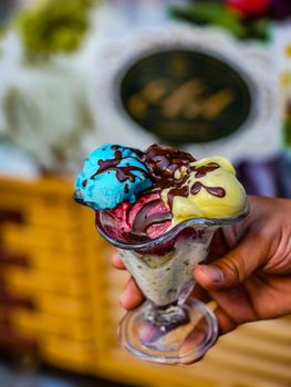 Close-up of a vibrant, multi-flavored ice cream sundae held in a glass, showcasing colorful scoops and chocolate drizzle.
