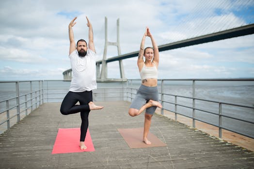 Two adults practicing yoga on mats outdoors with a bridge backdrop, showcasing healthy lifestyle.