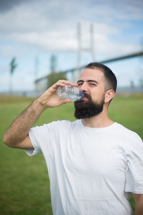 Free Portrait of a bearded man drinking water outdoors on a sunny day, bridge in the background. Stock Photo