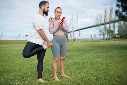 A couple practicing yoga in a park with a stunning bridge in Lisbon as a background, using a smartphone for guidance.