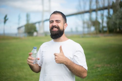 Free Cheerful man with a beard giving thumbs up while holding a water bottle in a park setting in Portugal. Stock Photo
