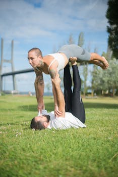 A couple performing acro yoga in a sunny park with a bridge in the background.