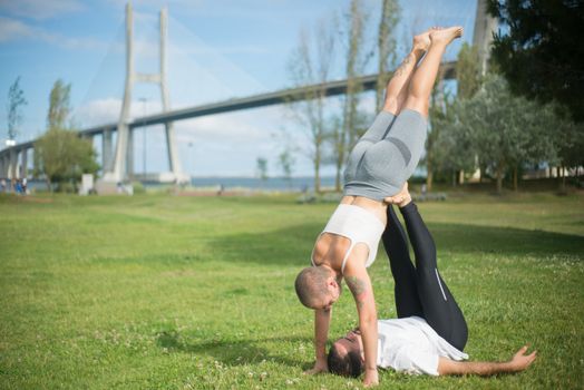 Two adults performing acro yoga outdoors with a bridge in the background.