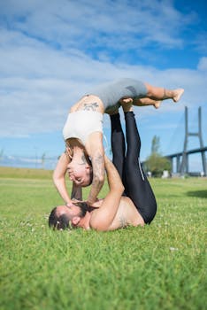 A couple performs an acro yoga pose on a green field under a clear sky near a bridge in Portugal.
