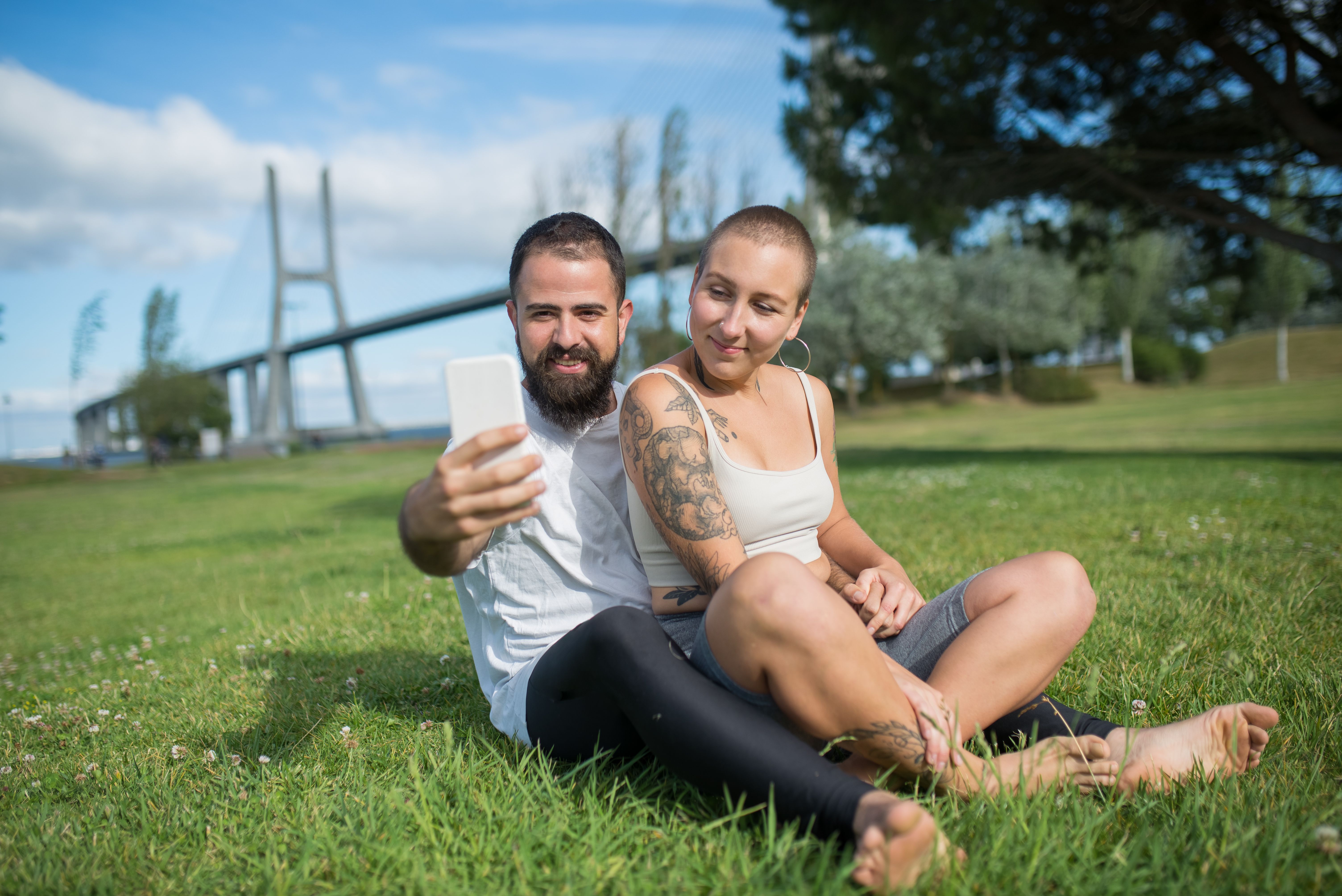 A Couple Sitting on Grass in the Park Taking a Selfie · Free Stock Photo