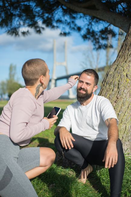 A joyful couple sitting under a tree exploring a smartphone in a sunny park setting.