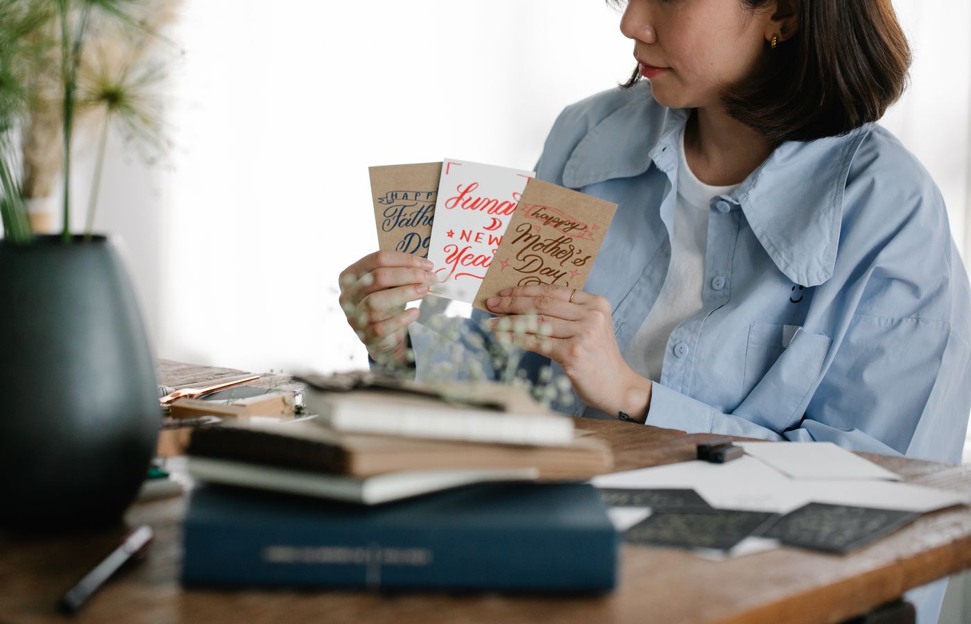 A woman selects various handmade greeting cards at a wooden table indoors, surrounded by books and plants.