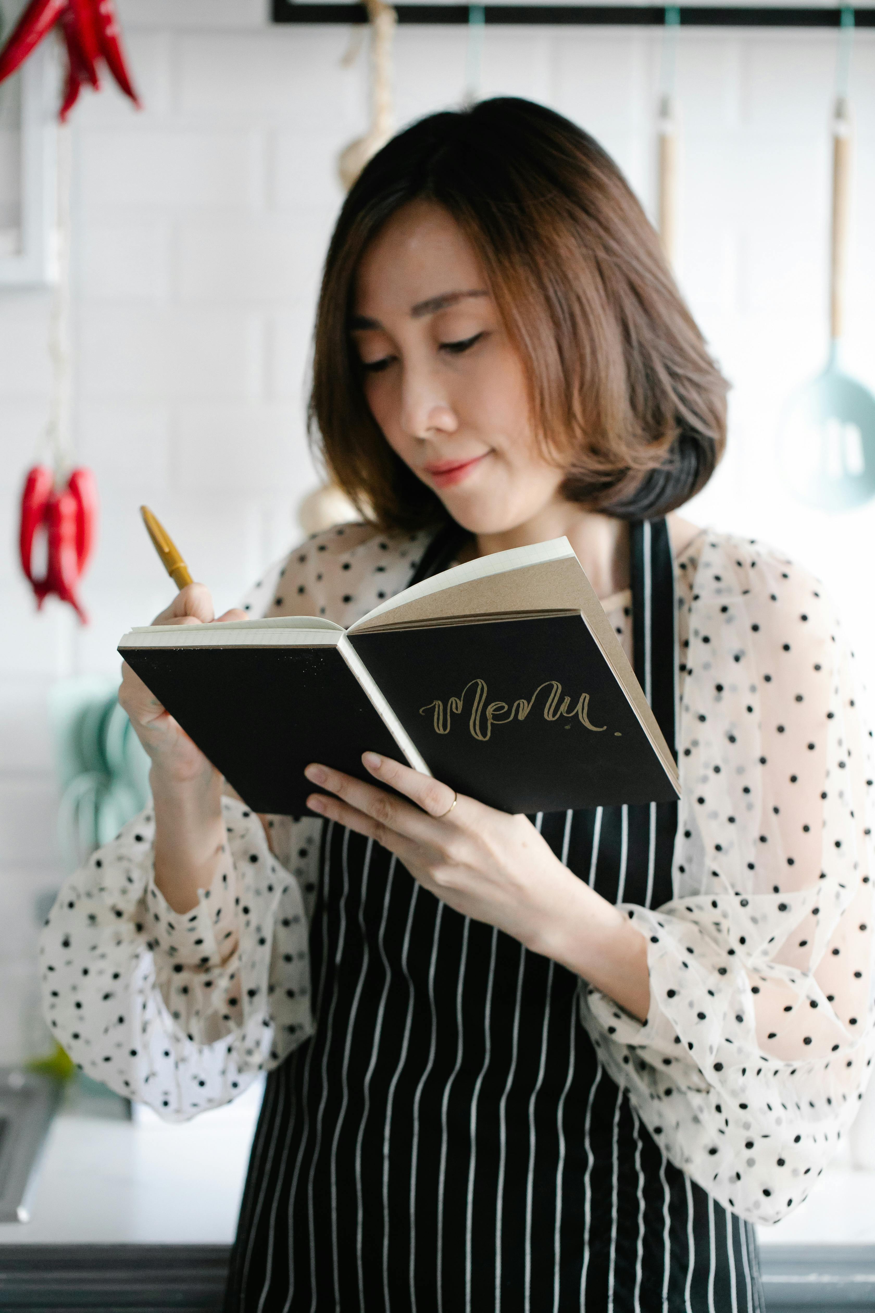 Asian woman in a kitchen writing in a menu book, wearing a polka dot blouse and apron.